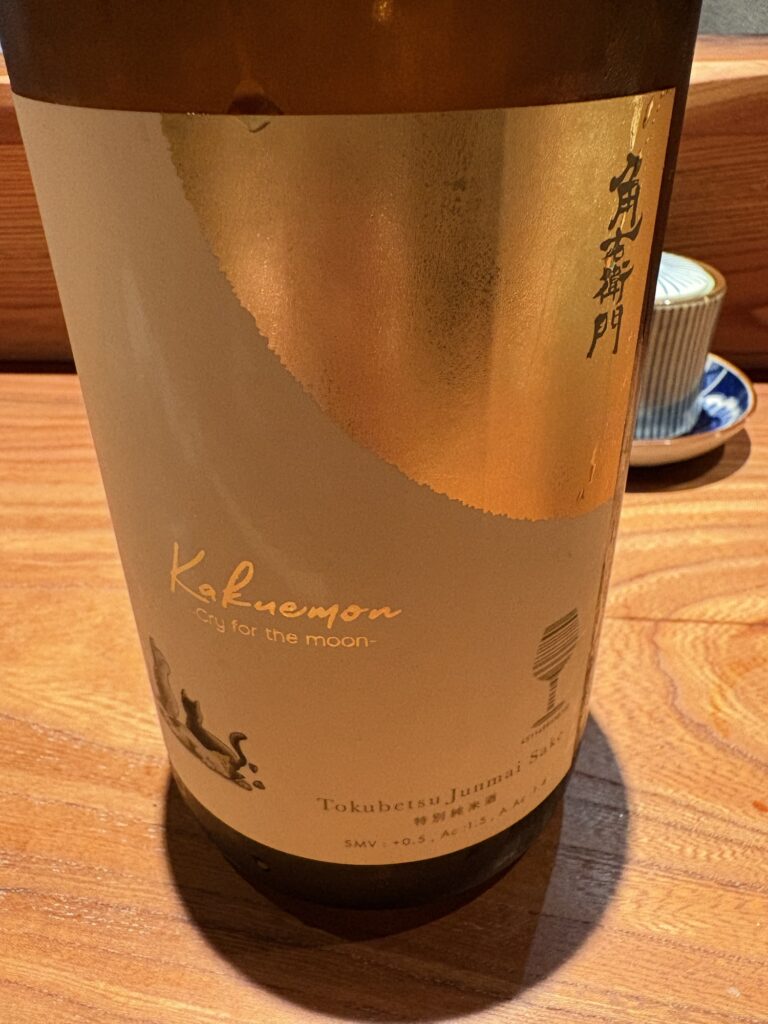 A bottle of Kakuenmon sake labeled 'Cry for the moon' with a textured gold and white design, sitting on a wooden surface.